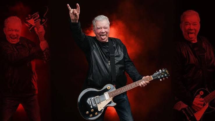 William Shatner sticks his tongue out while holding a black electric guitar and signaling devil horns with his hand.