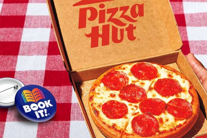 A personal pan pizza sits in a red box on a checkered tablecloth next to a vintage Book It! pin.