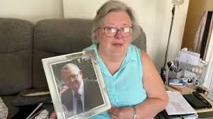 Catherine Curtis sits on a sofa, holding a framed photograph of her late husband, Michael Curtis.
