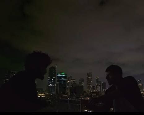 Silhouettes of two men on a Tel Aviv rooftop at night with a city skyline in the background.
