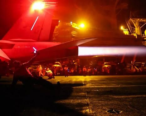 US military personnel stand on a darkened airfield near aircraft illuminated by red lighting.