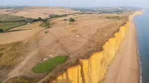 An aerial view of a dry golf course and fields with brown grass under a grey sky in Dorset, England.