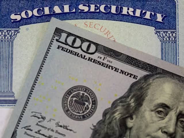 A close-up of a Social Security card on a wooden surface with a person holding a pen in the background.