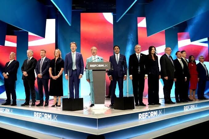 Nigel Farage singing the national anthem at the Reform UK conference surrounded by senior party officials.