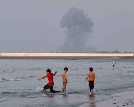 Three boys play in shallow water near Bandar Abbas as a plume of smoke rises from an explosion in the distance.