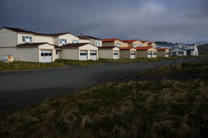 A dilapidated, abandoned Navy-built house on Adak island with peeling paint and broken windows during a clear summer day.