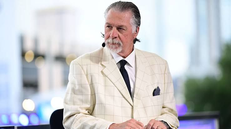 Barry Melrose, a former NHL coach and ESPN analyst, wearing a suit and smiling during a broadcast segment.