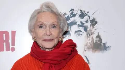 Dame Sian Phillips wearing a bright red jacket and a patterned scarf while speaking during an event.