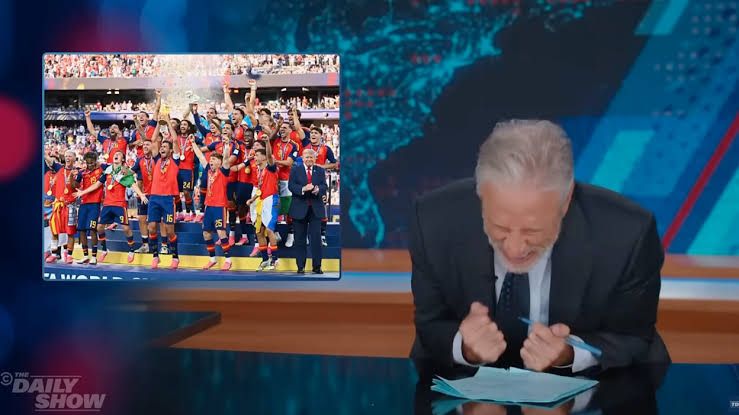 Jon Stewart hosting The Daily Show in front of an image showing the 2026 World Cup trophy presentation.