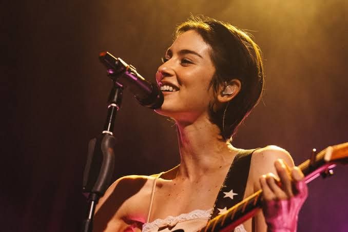Gracie Abrams performs with an acoustic guitar under red stage lights at the Bowery Ballroom in New York City.