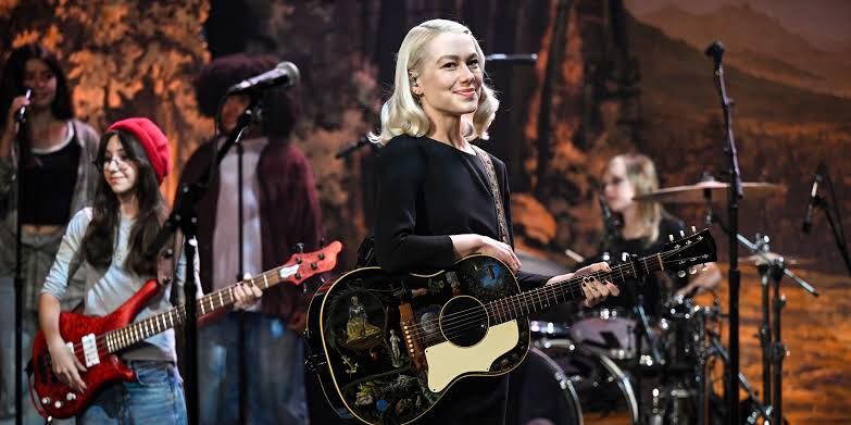 Phoebe Bridgers holding an electric guitar while standing on a stage with a microphone during a 2026 concert performance.