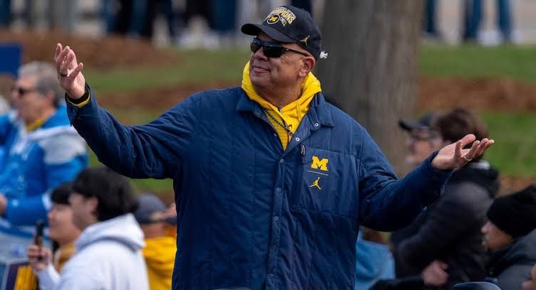 Warde Manuel standing on a football field wearing a Michigan windbreaker while speaking into a microphone.