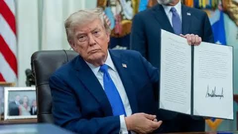 President Trump sits at the Oval Office desk while holding an executive order document for the camera.