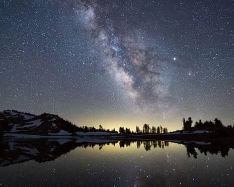 The Milky Way galaxy viewed above a dark lake at night with silhouetted mountain slopes in the background.