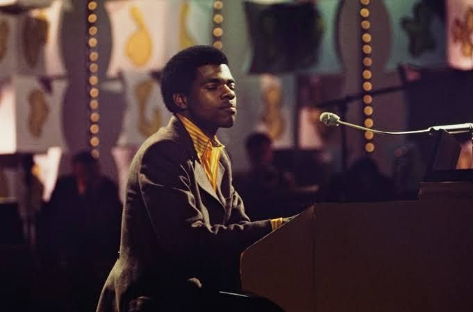 Billy Preston sitting at a keyboard next to George Harrison during a concert performance in 1974.