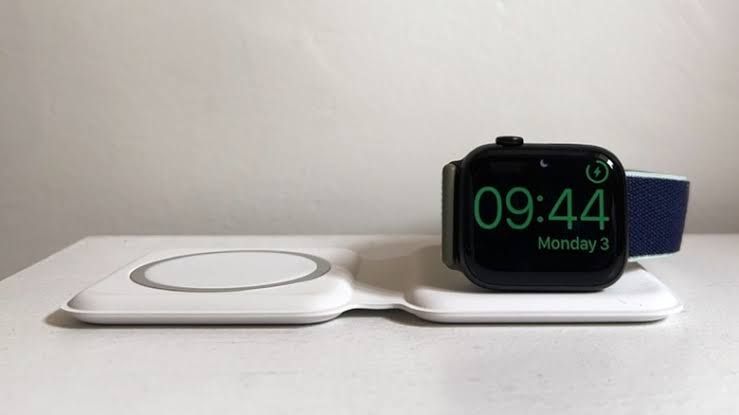 An Apple Watch charging on a white magnetic puck accessory next to a smartphone.