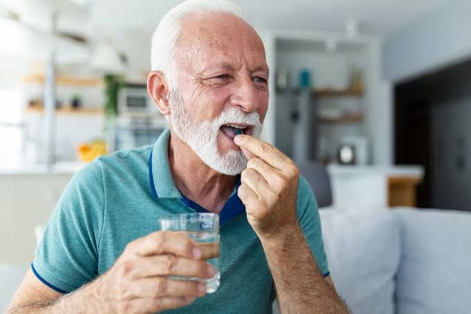 An older man taking a multivitamin pill with a glass of water in a well-lit home setting.
