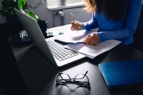 A woman at a desk in a home office reviewing tax documents and laptop paperwork.
