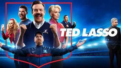 Jason Sudeikis as Ted Lasso standing on a soccer field wearing a blue tracksuit during filming for season four.
