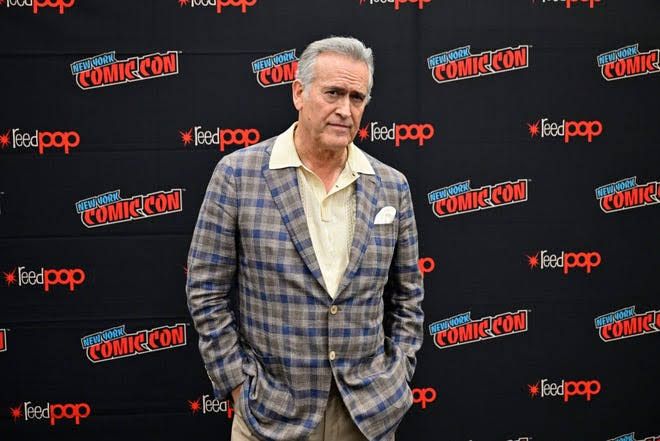 Bruce Campbell speaking at a press event in New York wearing a dark jacket.