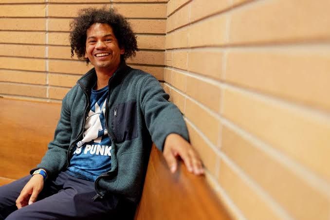 Mychal Threets stands in a library setting, smiling while holding a stack of books for his reading program.