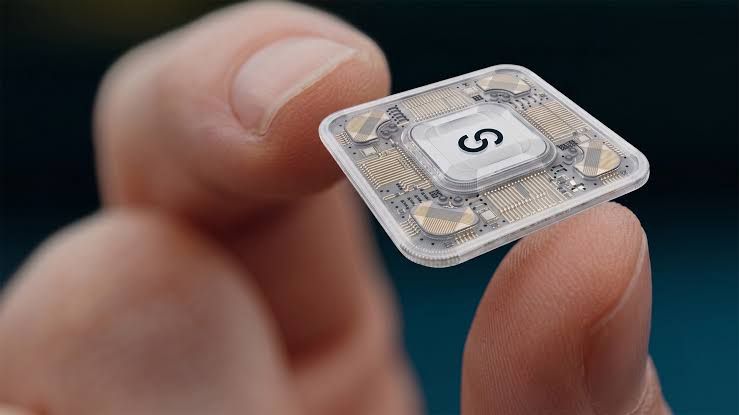 Close-up of a hand holding a small electronic circuit chip with the letter S on it against a dark background.