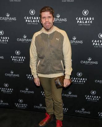 Perez Hilton stands on a red carpet while attending an industry event in a black suit.