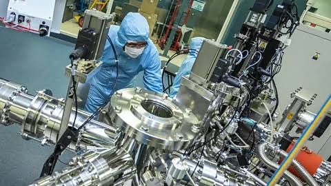 A researcher in a blue lab suit and mask inspects a cylindrical metal device in the CSIRO quantum fabrication laboratory.