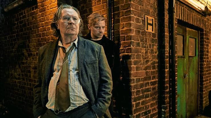 Gary Oldman in a trench coat standing in a dimly lit London alleyway for the new season of the show Slow Horses.