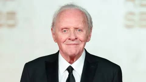 Sir Anthony Hopkins smiling in a music studio while wearing a blue scarf and blazer.