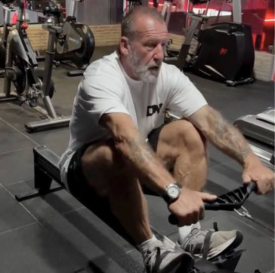 Dorian Yates sitting on a rowing machine during an intense workout session to maintain his physical conditioning.
