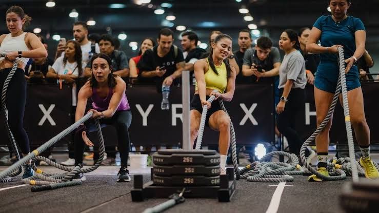 Athletes performing a sled push maneuver on a turf track during a Hyrox fitness competition in an indoor arena.