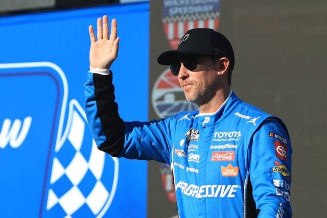 Denny Hamlin in his racing suit standing by his vehicle at the Indianapolis Motor Speedway during the Brickyard 400.