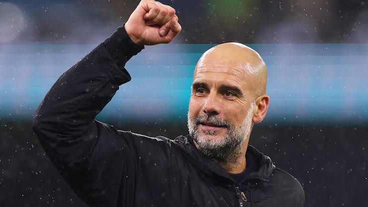 Pep Guardiola in a dark suit standing on a pitch, acknowledging the crowd after a Manchester City victory.