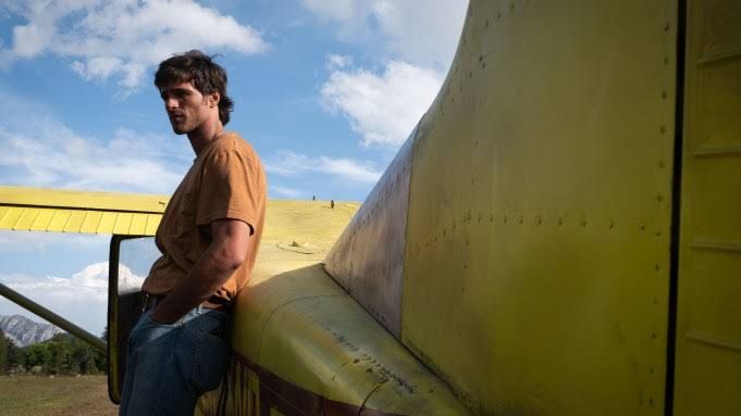 Jacob Elordi as the pilot Hig standing in front of a yellow Cessna plane in a post-apocalyptic mountain landscape.