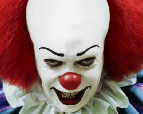 Tim Curry as Pennywise the clown with white face paint and a sinister grin.
