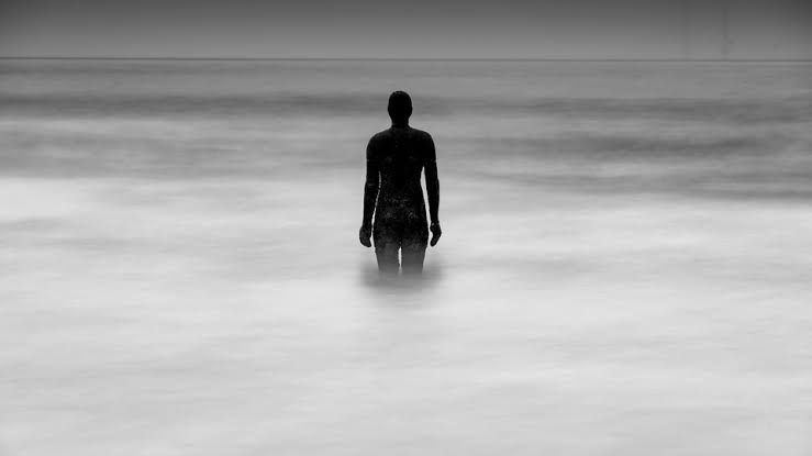 A solitary statue stands in shallow water against a foggy, grey horizon, symbolizing existential isolation.