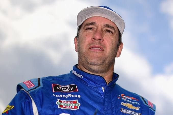 Elliott Sadler wearing a racing uniform stands next to a Kaulig Racing truck at a speedway.