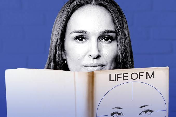 Natalie Portman holding a copy of the book Life of M against a blue background.