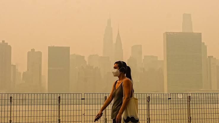 A woman walks through a smoke-filled park in New York City with an orange haze visible in the background.