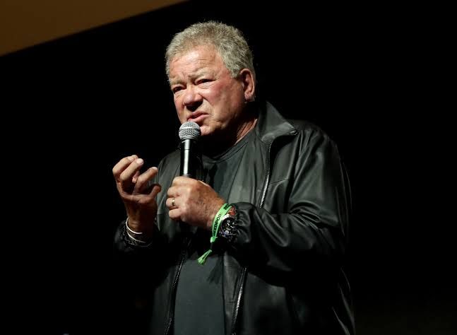 William Shatner speaking at a conference microphone during a public event.