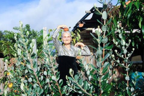 A smiling woman with grey hair holds a persimmon above her head while standing in a garden.