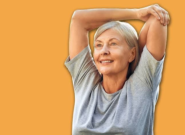 A woman over 65 practicing a sit-to-stand exercise using a sturdy chair in her living room to improve leg strength.