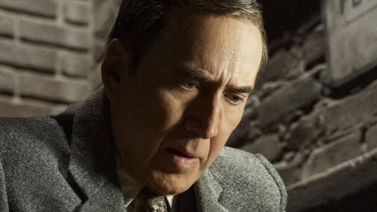 Nicolas Cage stands in a dimly lit, moody set portraying the noir private investigator character.