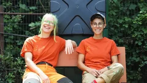 Steph Robinson and ffin Jordao in orange work uniforms sitting on a bench at a community composting site.