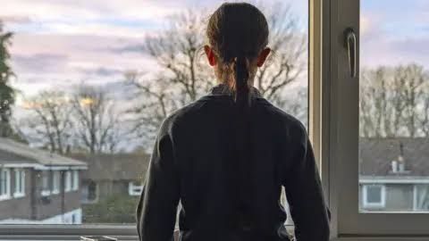 A young girl looks out of a window as medical professionals evaluate systemic mental health gaps for children in England.