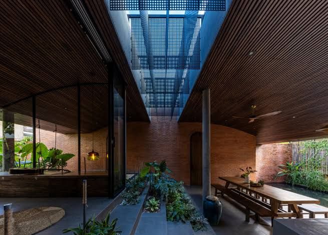 An open living space featuring wood ceilings and brick walls with indoor plants and a pond reflecting natural light.