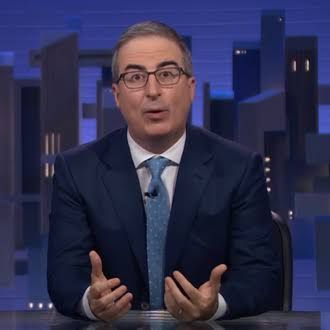 John Oliver sitting at his desk on the set of Last Week Tonight with the HBO logo visible.