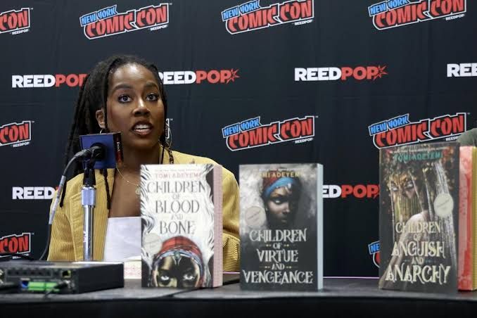 Tomi Adeyemi speaking at a public event with her books displayed on a table.