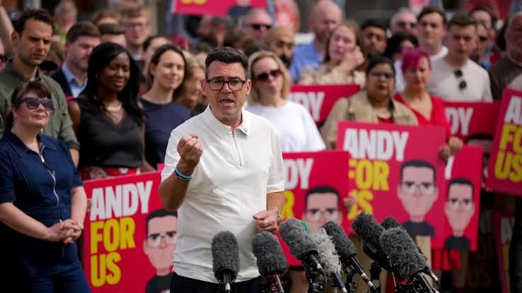 Andy Burnham walking through a revitalized Manchester city center past modern office buildings and bustling public spaces.